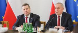 Minister Czarnek i mężczyzna w garniturze siedzą za stołem, przed nimi kilka zielonych butelek z wodą, mikrofony, za nimi flagi Polski i UE.