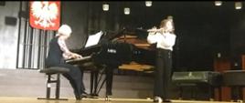 Na zdjęciu widoczna jest scena w sali koncertowej. Na pierwszym planie występują dwie osoby – jedna gra na fortepianie, a druga na flecie poprzecznym.
Pianistka to starsza kobieta z krótkimi, jasnymi włosami. Siedzi na czarnej ławie i gra na fortepianie marki Fazoli. Jest ubrana w ciemny strój i ma wzorzystą bluzkę.
Flecistka to młoda kobieta w białej bluzce i czarnych spodniach. Stoi po prawej stronie sceny, trzymając flet poprzeczny przy ustach.
W tle znajduje się godło Polski – biały orzeł na czerwonym tle. Scena jest dobrze oświetlona, a w tle widać także inne instrumenty, w tym fortepian i pianino. Całość wskazuje na recital lub konkurs muzyczny.