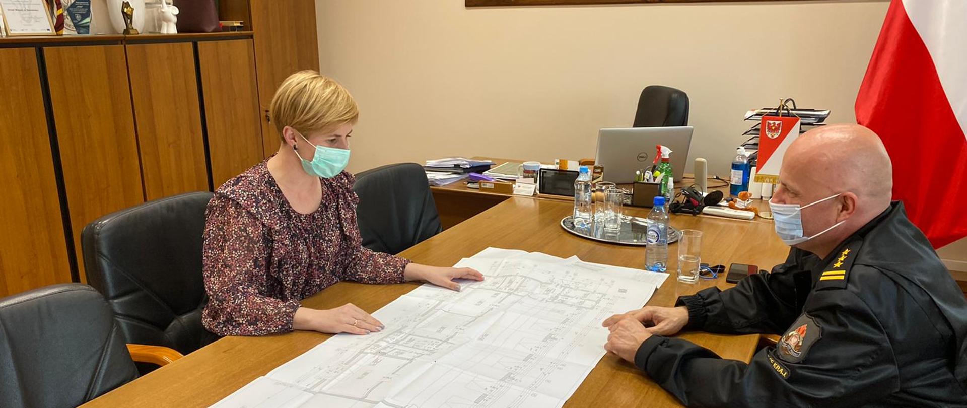 Fotografia przedstawia ujęcie gabinetu Burmistrz Drezdenka. Od lewego dolnego narożnika kadru ciągnie się stół konferencyjny zakończony biurkiem Burmistrz. Przy stole konferencyjnym po jego obu stronach siedzą: po prawej stronie Komendant Powiatowy PSP w Strzelcach Krajeńskich – starszy brygadier Tomasz Górniak w mundurze dowódczo-sztabowym z założoną maseczką chirurgiczną; po lewej stronie Burmistrz Drezdenka – Pani Karolina Piotrowska, z założoną maseczką chirurgiczną. Na stole konferencyjnym rozłożony jest rzut kondygnacji projektu budowlanego. Za biurkiem, na ścianie widoczny duży rysunek panoramy Drezdenka z przed kilku wieków oprawiony w drewnianą ramę. Obok obrazu po prawej stronie kadru widoczna stojąca flaga Polski. Po lewej stronie kadru pod ścianą stoją meble, powyżej mebli widoczny oprawiony w ramę herb Drezdenka.