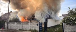 Budynek mieszkalny z którego wydobywają się kłęby dymu oraz ogień. Przed budynkiem betonowe ogrodzenie i metalowa brama. Obok drugi budynek mieszkalny.