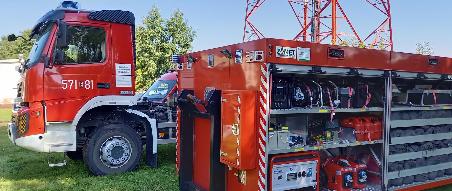 Zdjęcie przedstawia stojący w tle czerwony samochód specjalny o numerach operacyjnych 571-81. Na pierwszym planie na trawie stoi kontener kwaterunkowy z otwartymi skrytkami. Wewnątrz widoczne wyposażenie kontenera m.in. agregaty, przedłużacze, śpiwory.