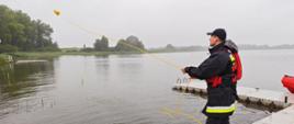 Na zdjęciu widoczny jest strażak w czarno-czerwonym stroju ratowniczym i kapoku, stojący na drewnianym pomoście nad jeziorem. Mężczyzna rzuca żółtą linę ratowniczą z przymocowanym pływakiem w stronę wody. Na pierwszym planie widać zwiniętą część liny leżącą na pomoście. W tle jezioro otoczone jest roślinnością i drzewami, a na horyzoncie widać zamglony, pochmurny krajobraz. Pogoda jest deszczowa, co sugeruje mokrą nawierzchnię i trudne warunki.