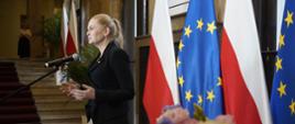 Zdjęcie z boku, minister Nowacka stoi i mówi do mikrofonu na stojaku, za nią przy ścianie rząd flag Polski i UE.