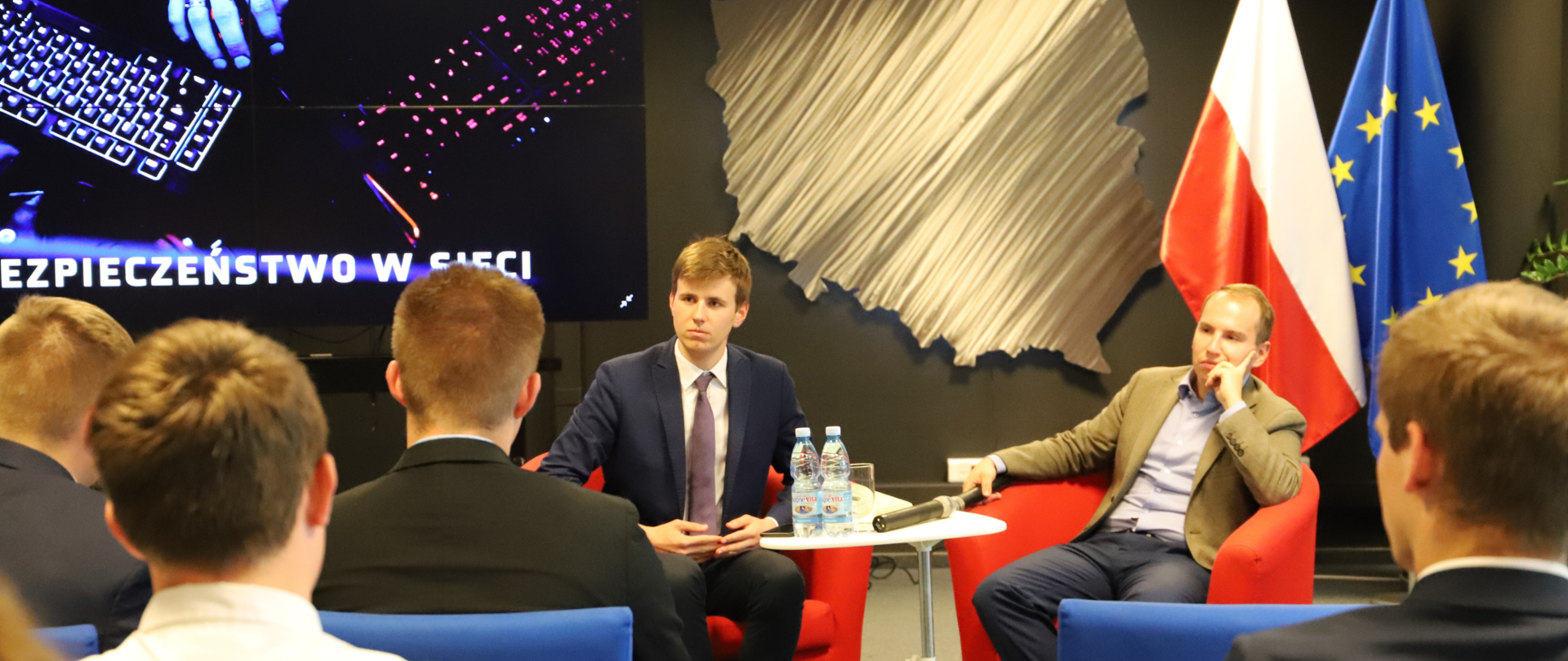 Sala konferencyjna. Kilka osób siedzi na wprost stolika, przy którym siedzi dwóch mężczyzn.