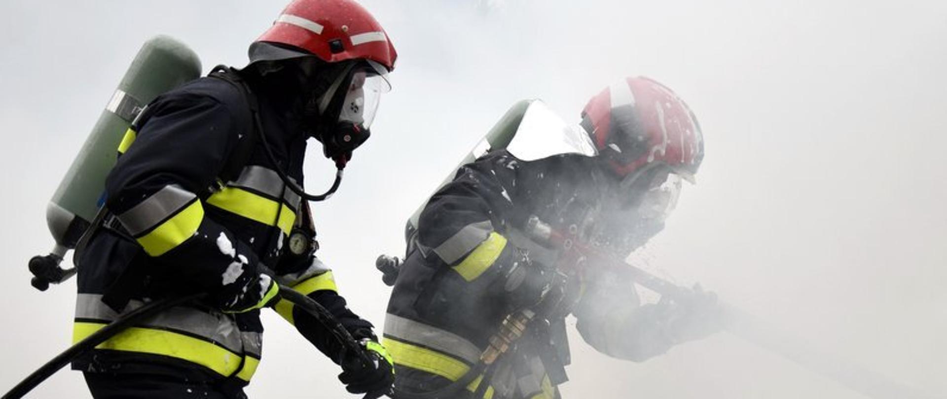 Dwóch strażaków ubranych w ubrania specjalne (nomexy) w hełmach i aparatach powietrznych stoi w zadymianym pomieszczeniu, gasząc niewidoczny na zdjęciu ogień