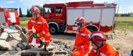 Na zdjęciu trzech strażaków w pomarańczowych kombinezonach. W tle wóz strażacki. Strażacy skupieni na ćwiczeniu. W centralnym miejscu zdjęcia skrzynka ze sprzętem.