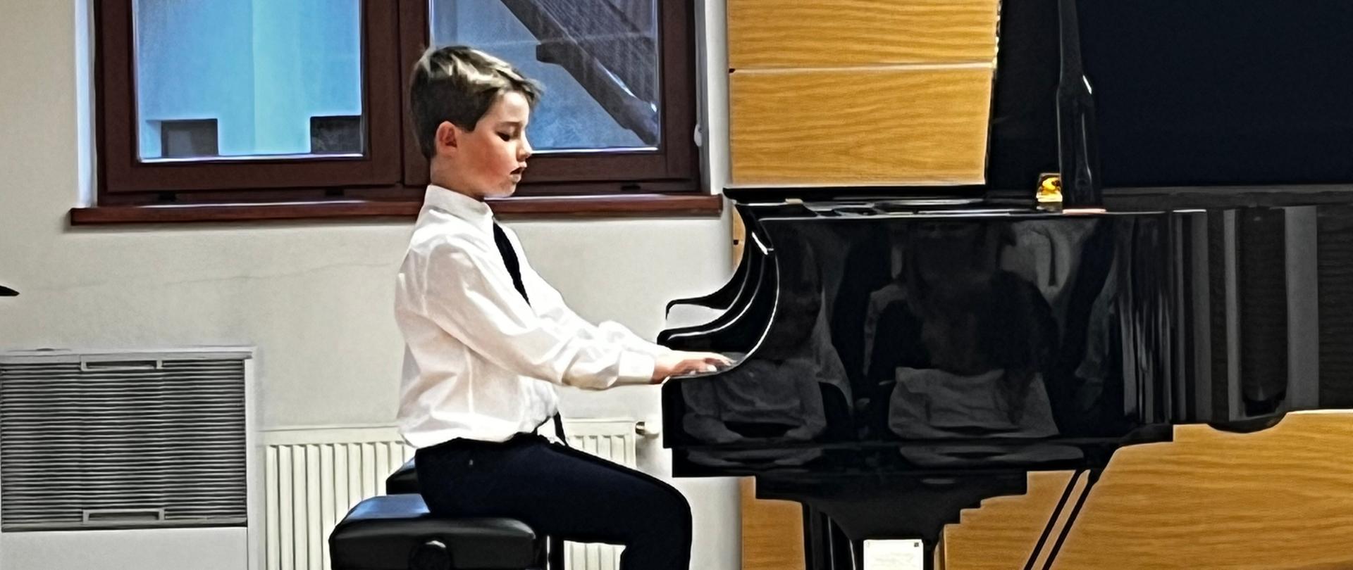 Zdjęcie przedstawia młodego chłopca, prawdopodobnie ucznia, podczas występu na koncercie lub audycji muzycznej, grającego na fortepianie. Centralny punkt: Młody chłopiec (szacowany wiek szkolny), siedzi na czarnej ławie fortepianowej, grając na dużym, czarnym fortepianie koncertowym. Chłopiec jest ubrany w białą koszulę i ciemny krawat lub muszkę oraz ciemne spodnie. Ma krótkie, jasne włosy. Jego uwaga jest skupiona na instrumencie. Pod jego stopami znajduje się regulowany podnóżek, typowy dla młodych pianistów. Fortepian (półkoncertowy lub koncertowy) jest lśniąco czarny, z otwartą klapą, co wskazuje na akustykę sali. Scena/Sala ma drewniane panele na prawej ścianie, które dodają ciepła. Po lewej stronie widać duże okno z żaluzjami lub zasłoną w kolorze musztardowym/żółtym. Przez szybę widać fragment budynku za oknem. Pod oknem stoi biały grzejnik żeberkowy oraz mały, przenośny klimatyzator lub oczyszczacz powietrza.W prawym dolnym rogu widoczny jest fragment głowy innej osoby (dziewczynki) z fioletową kokardą, co sugeruje, że publiczność jest blisko sceny.