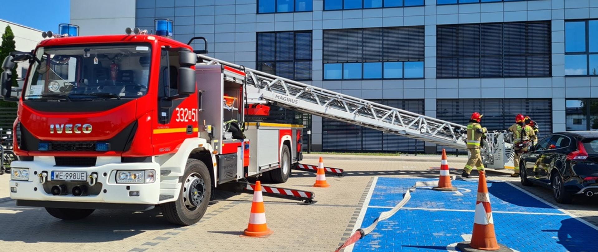 Na zdjęciu widać na pierwszym planie widać pojazd specjalny drabina pożarnicza. Pojazd w kolorze czerwonym ze srebrnymi i białymi elementami zabudowy. Pojazd posiada rozłożony w stronę ziemi park drabinowy zakończony koszem. Przy koszu znajduje się trzech strażaków ubranych w ubrania specjalne w kolorze żółtym. Na głowach mają założone czerwone hełmy. Dwóch z nich na plechach posiada założone również aparaty ochrony dróg oddechowych. Przy pojeździe na ziemi znajduje się również rozłożona linia gaśnicza. W tyle za pojazdem widać zakład produkcyjny w którym odbywają się ćwiczenia.