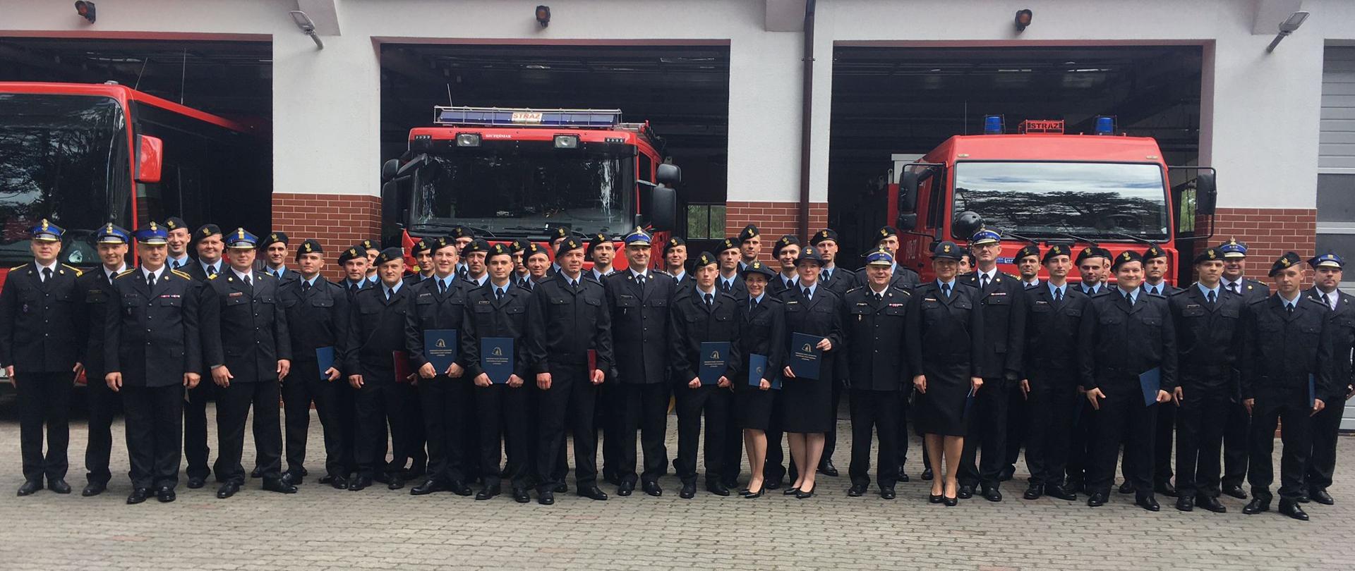 Na zdjęciu znajdują się strażacy kończący kurs podstawowy, wraz z kadrą naukową oraz zaproszonymi przedstawicielami KW PSP w Szczecinie oraz SA PSP w Poznaniu. w tle widoczne są pojazdy strażackie oraz budynek jednostki ratowniczo-gaśniczej wojewódzkiego ośrodka szkolenia.