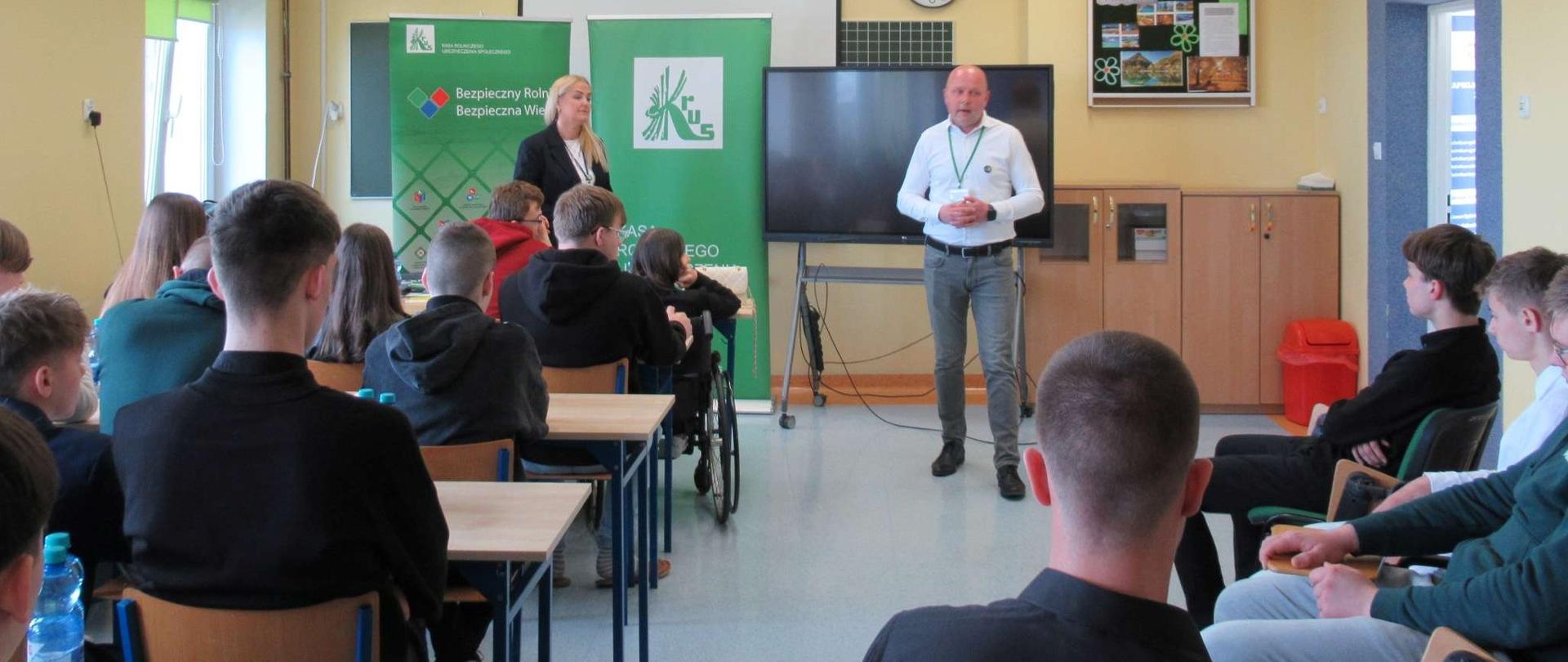 Mężczyzna i kobieta prowadzą prezentację w klasie szkolnej przed grupą uczniów siedzących w ławkach, za nimi stoją zielone roll-upy i ekran multimedialny.