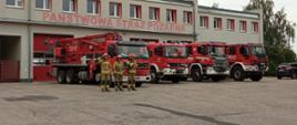 Zdjęcie przedstawia 4 ciężarowe samochody strażackie stojące przed obiektem jednostki z włączonymi sygnałami świetlnymi. Na ich tle 3 strażaków ubranych w piaskowe mundury specjalne stoi w rzędzie.