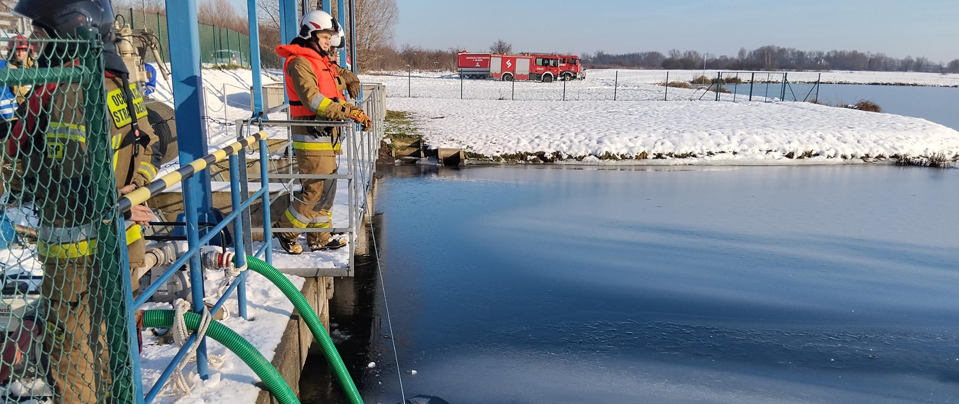 Na zdjęciu widać węże ssawne zanurzone w zbiorniku retencyjnym. Na platformie nad zbiornikiem stoją strażacy nadzorujący pracę pomp pożarniczych. Strażacy wyposażeni są w kamizelki asekuracyjne.