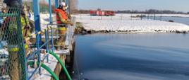 Na zdjęciu widać węże ssawne zanurzone w zbiorniku retencyjnym. Na platformie nad zbiornikiem stoją strażacy nadzorujący pracę pomp pożarniczych. Strażacy wyposażeni są w kamizelki asekuracyjne.