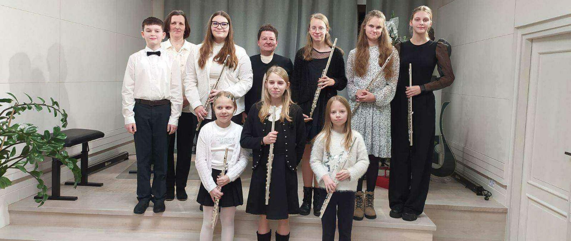 Zdjęcie przedstawia grupę uczniów i nauczycieli podczas koncertu klasy fletu. Dziewięć z dziesięciu osób trzyma flety poprzeczne, a wszyscy są odświętnie ubrani. Grupa stoi na podium w jasnym, nowoczesnym wnętrzu. W tle znajduje się zielona choinka z białymi ozdobami i prezentami. W rogu pomieszczenia widoczny jest dekoracyjny klucz wiolinowy oraz popiersie patrona.