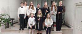Zdjęcie przedstawia grupę uczniów i nauczycieli podczas koncertu klasy fletu. Dziewięć z dziesięciu osób trzyma flety poprzeczne, a wszyscy są odświętnie ubrani. Grupa stoi na podium w jasnym, nowoczesnym wnętrzu. W tle znajduje się zielona choinka z białymi ozdobami i prezentami. W rogu pomieszczenia widoczny jest dekoracyjny klucz wiolinowy oraz popiersie patrona.