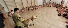 Zdjęcie przedstawia spotkanie dzieci z psem ratownikiem i jego opiekunem na szkolnej sali gimnastycznej 