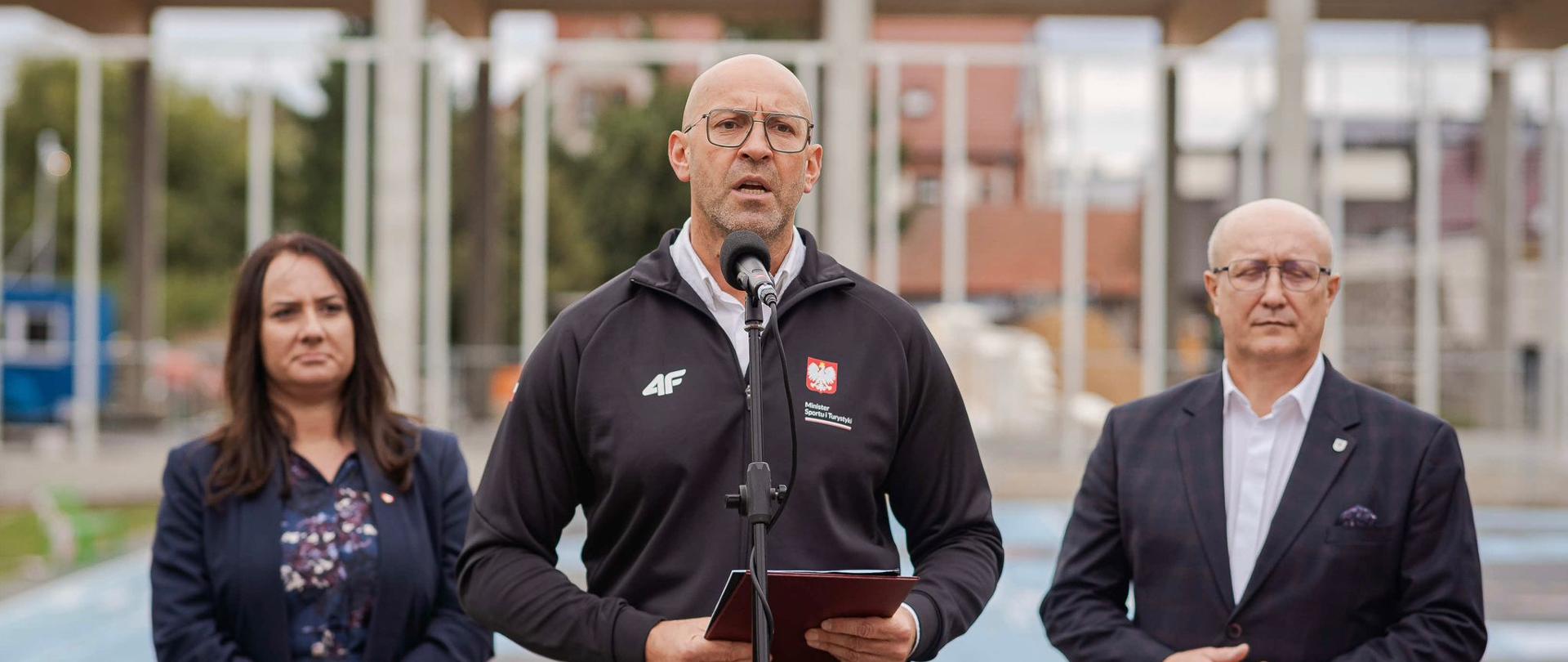 Zdjęcie z konferencji prasowej. Na środku przy mikrofonie stoi minister sportu i Turystyki Jakub Rutnicki. Po bokach, nieco z tyłu, stoi dwoje innych uczestników konferencji. W tle, niewyraźnie, widać budowę hali sportowej.