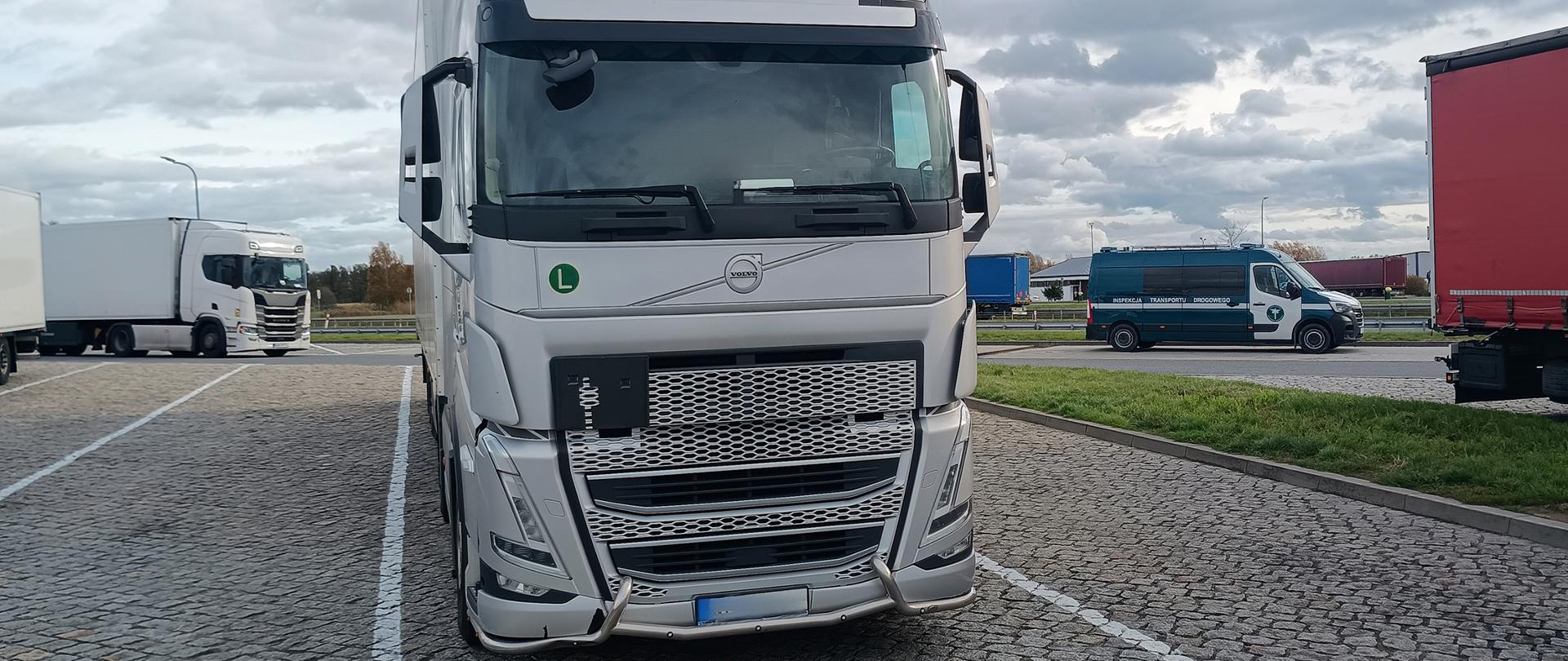 Biały ciągnik siodłowy marki Volvo zaparkowany na parkingu z kostki brukowej. W tle po lewej stronie znajdują się inne ciężarówki, a po prawej częściowo widoczna naczepa z czerwoną plandeką. W dalszym planie po prawej stronie stoi furgon z napisem Inspekcja Transportu Drogowego.