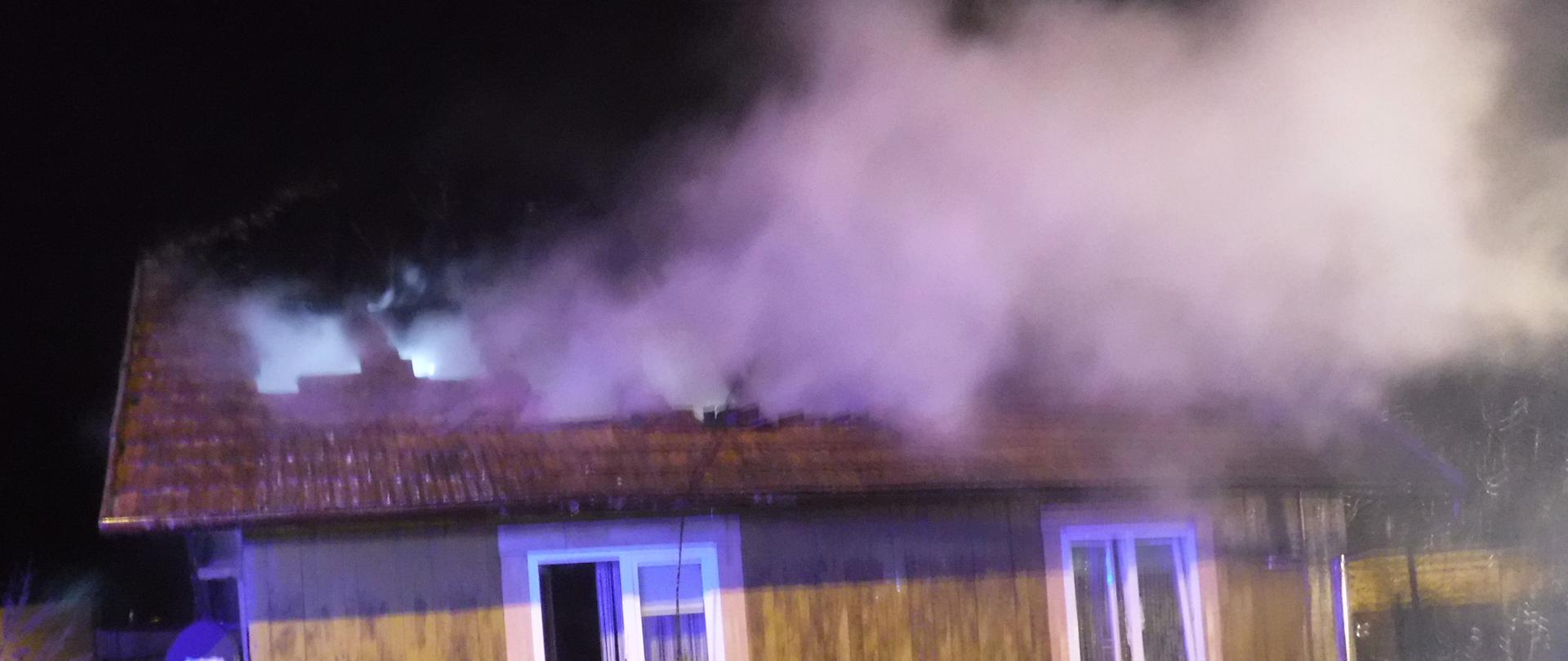 Zdjęci przedstawia drewniany budynek mieszkalny, w którym doszło do pożaru. Można zaobserwować znaczące braki w pokryciu dachowym. Na zdjęciu możemy zaobserwować parę wodną zmieszaną z dymem unoszącą się nad budynkiem. Widać wąż strażacki poprzez, który przepływa woda w celach gaśniczych. Zauważamy również ogrodzenie budynku. Dwa okna w kolorze białym na zdjęciu są otwarte. Z lewej strony budynku możemy zauważyć zniszczone elementy konstrukcji dachu. 