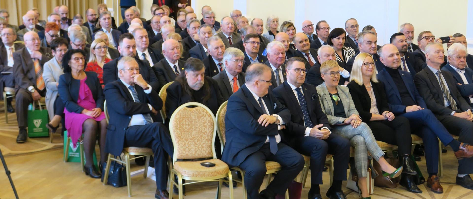 Na sali na ustawionych w rzędy krzesłach siedzi dużo ludzi, w pierwszym rzędzie między mężczyznami w garniturach siedzi wiceminister Mrówczyńska.