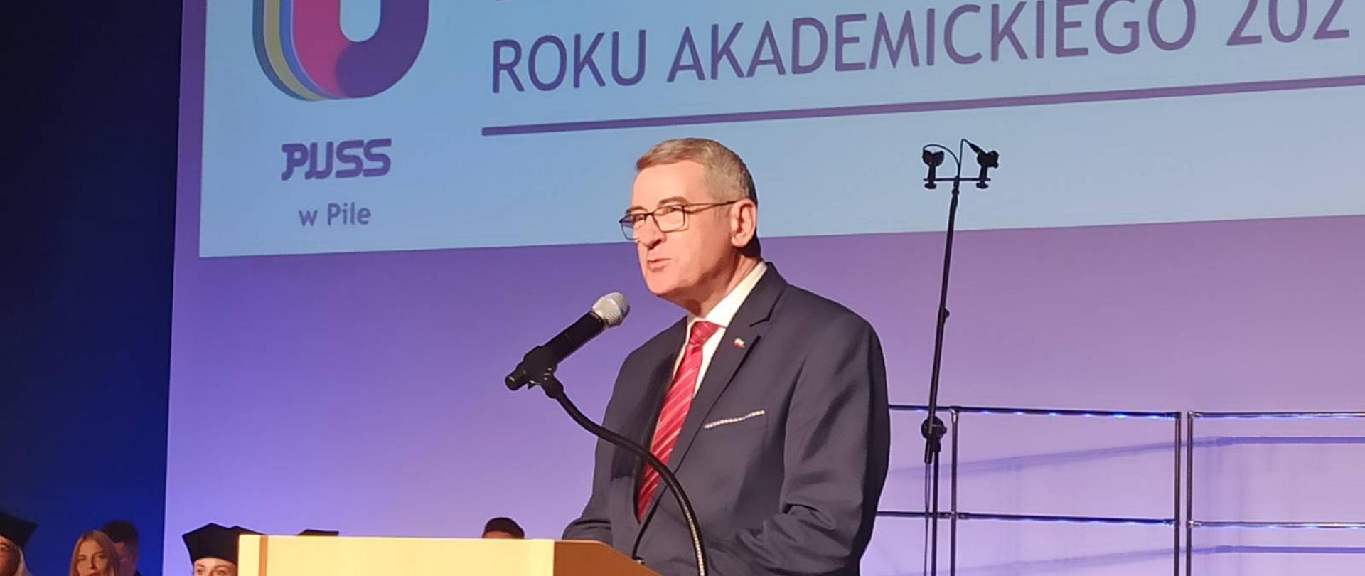 Państwowa Uczelnia im. Stanisława Staszica zainaugurowała rok akademicki 2021/2022 wiceminister przemawia do mikrofonu, w tle plansza z napisem inauguracja roku akademickiego. Z lewej strony słuchacze. 