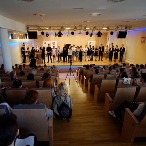 zdjęcie przedstawia maturzystów stojących w szeregu na scenie sali koncertowej, widok z tyłu widowni sali koncertowej
