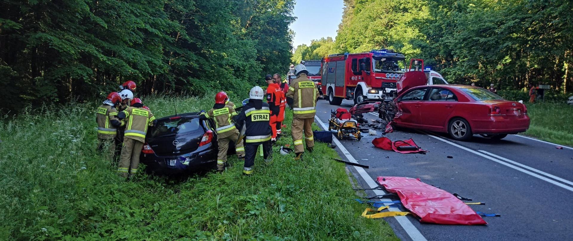 Zdjęcie przedstawia wypadek dwóch samochodów osobowych oraz współprace służb ratowniczych straży pożarnej oraz pogotowia ratunkowego podczas wydobywania osoby poszkodowanej z wraku pojazdu.