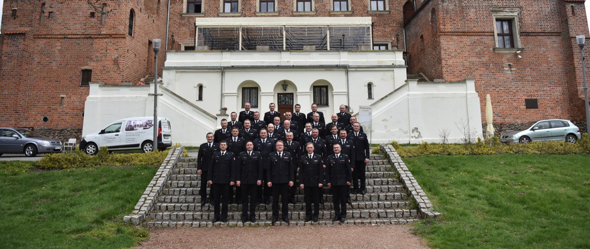 na zdjęciu grupa strażaków w mundurach galowych stojących na schodach prowadzących do zamku, w tle część zamku 