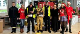 Na zdjęciu widzimy grupę ośmiu osób zaangażowanych w akcję charytatywną „Szlachetna Paczka”. W centralnej części fotografii stoją przedstawiciele służb mundurowych: strażak w charakterystycznym piaskowym mundurze oraz dwaj strażnicy miejscy ubrani w granatowe mundury i jaskrawe, żółte kamizelki odblaskowe. Mężczyźni trzymają w dłoniach dyplomy lub plakaty potwierdzające udział w inicjatywie. Po obu stronach funkcjonariuszy stoją wolontariusze, z których część ma na sobie czerwone koszulki z logotypem akcji i hasłem „Człowiek pełen dobra”. Świąteczną atmosferę podkreśla kobieta w czapce świętego Mikołaja oraz tło udekorowane bożonarodzeniowymi akcentami.
Scena rozgrywa się w jasnym pomieszczeniu, które wygląda na szkołę lub placówkę publiczną. W tle, za plecami uczestników, widoczna jest dekoracja z motywem serca oraz liczne prezenty zapakowane w kolorowy papier i folię, które czekają na przekazanie potrzebującym. Grupa prezentuje się z dumą i uśmiechem, co podkreśla wspólnotowy charakter wydarzenia oraz współpracę lokalnych służb z wolontariuszami na rzecz pomocy innym. Całość kompozycji emanuje pozytywną energią i duchem bezinteresownego wsparcia.

