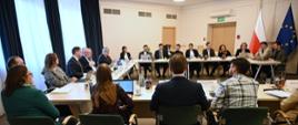 Na zdjęciu widoczni są uczestnicy spotkania Ministerstwa Klimatu i Środowiska z przedstawicielami Międzynarodowej Agencji Energii. Uczestnicy wydarzenia siedzą przy stołach ustawionych w kształt litery U. Po lewej stronie, w rogu sali widoczne są dwie flagi - flaga Unii Europejskiej i flaga Polski. 