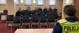 W pomieszczeniu na krzesłach siedzi kilkunastu strażaków w czarnych mundurach. Przed nimi policjantka w odblaskowej kamizelce.