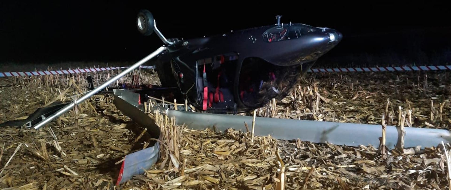 Przewrócony wiatrakowiec leży na polu po skoszonej kukurydzy podwozie znajduje się na górze, połamany wirnik główny na dole, widać uszkodzenia szyb kabiny i urwane jedno z kół podwozia, w tle taśma ostrzegawcza wyznaczająca miejsce zdarzenia.