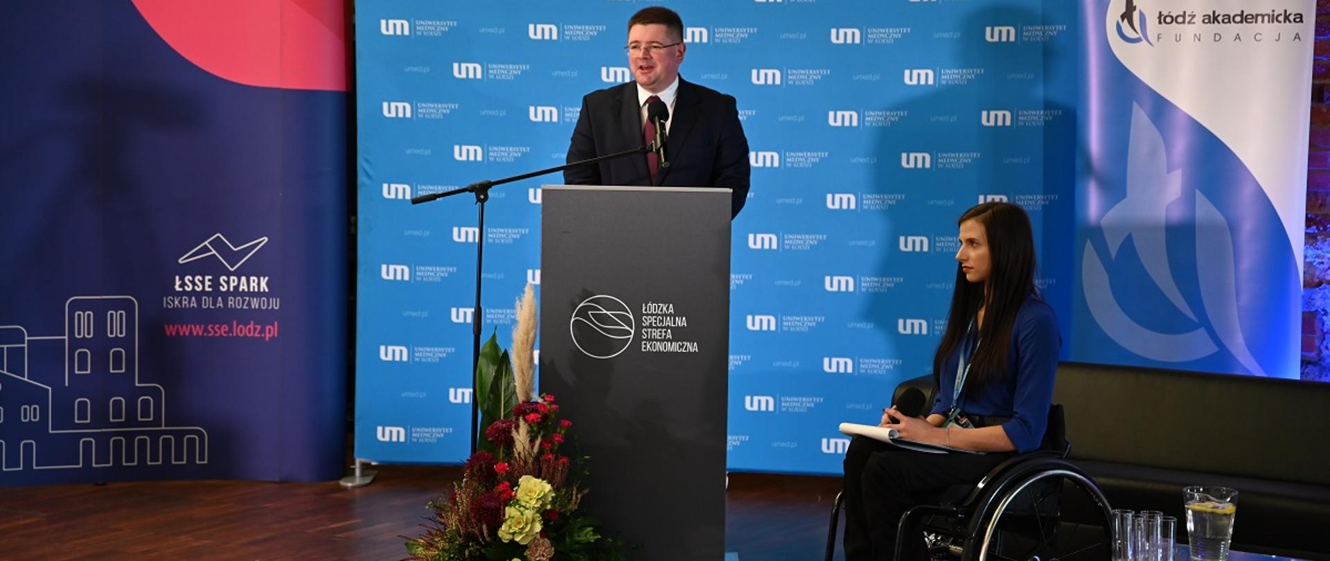 Wiceminister Tomasz Rzymkowski uczestnikiem konferencji „(Nie)pełnosprawni na rynku pracy”, Wiceminister Tomasz Rzymkowski przemawia podczas konferencji, stoi przy mównicy, obok na wózku inwalidzkim siedzi osoba niepełnosprawna.