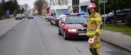 Ulica Kamińskiego w Rawiczu. Na jezdni stoi strażak w ubraniu specjalnym w kolorze piaskowym i w czerwonym hełmie. W rękach trzyma liazak do kierowania ruchem oraz radiotelefon. Za nim zaparkowany jest samochód operacyjny straży pożarnej oraz karetka pogotowia. W tle poruszające sie pojazdy. 