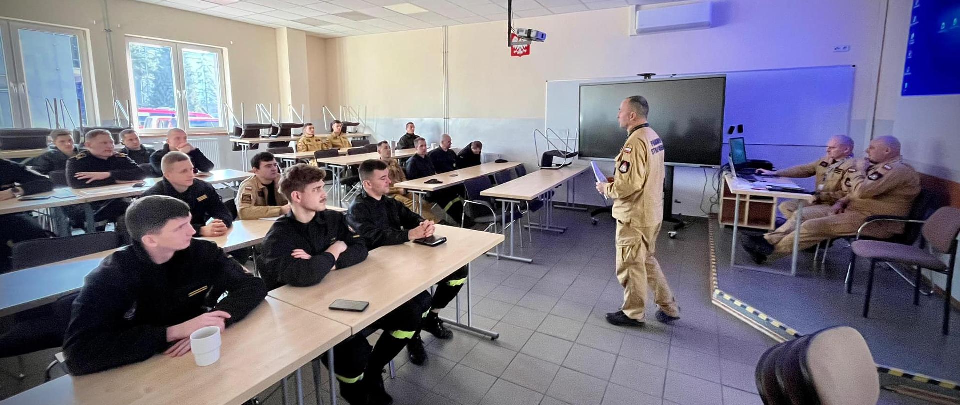 Z przyjemnością opiszę to zdjęcie.
**Zdjęcie 8b8b4c11-12a0-4a00-bce9-79ba64c04261.jpg:**
Zdjęcie przedstawia salę lekcyjną lub szkoleniową, w której odbywają się zajęcia dla grupy strażaków. W centralnej części kadru, odwrócony plecami do widza, stoi instruktor w beżowym mundurze strażackim, trzymający w rękach dokumenty i przemawiający do zebranych. Przed nim, przy długich stołach, siedzi kilkunastu strażaków w różnych typach uniformów (zarówno beżowych, jak i ciemnych, prawdopodobnie czarnych), uważnie słuchających wykładu. Na końcu stołów, po prawej stronie, siedzi dwóch starszych mężczyzn w beżowych mundurach, również słuchających. W tle, za instruktorem, znajduje się duży monitor lub tablica interaktywna, a obok niego okna, przez które wpada dzienne światło. Sala jest dobrze oświetlona, a na suficie widoczne są lampy fluorescencyjne. Atmosfera na zdjęciu jest skupiona i edukacyjna.