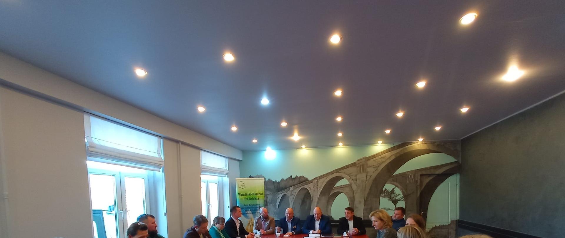 Grupa osób uczestniczących w spotkaniu siedzących przy stole. W tle okna, ściana z namalowanymi
mostami i baner Warmińsko-Mazurskiej Izby Rolniczej.