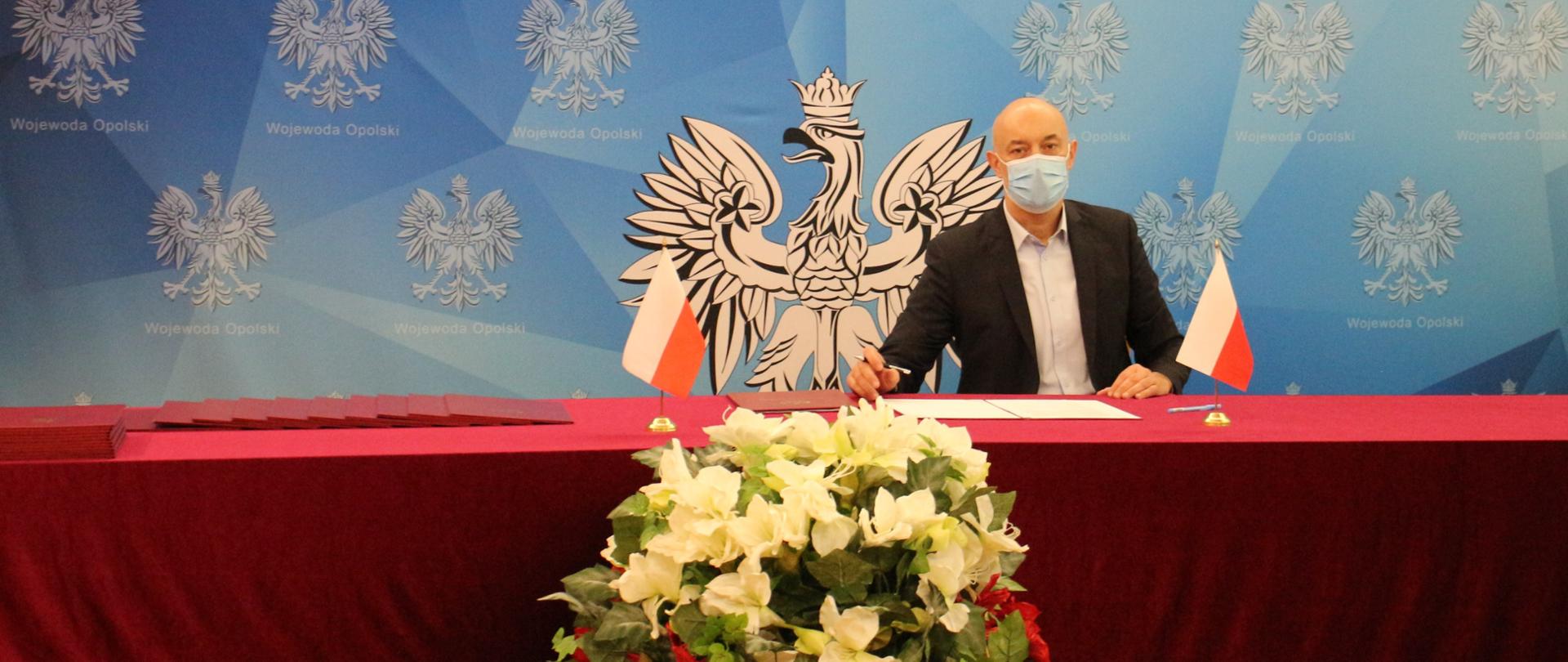 wojewoda opolski Adrian Czubak siedzi za stołem, przed nim na stole leżą dokumenty, z nim niebieska ścianka tekstylna z godłem