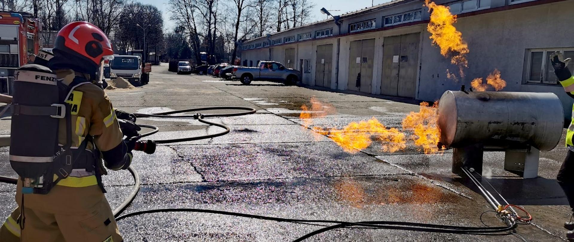 Po lewej dwóch ćwiczących strażaków w ubraniach bojowych z butlą, po prawej palący się zbiornik