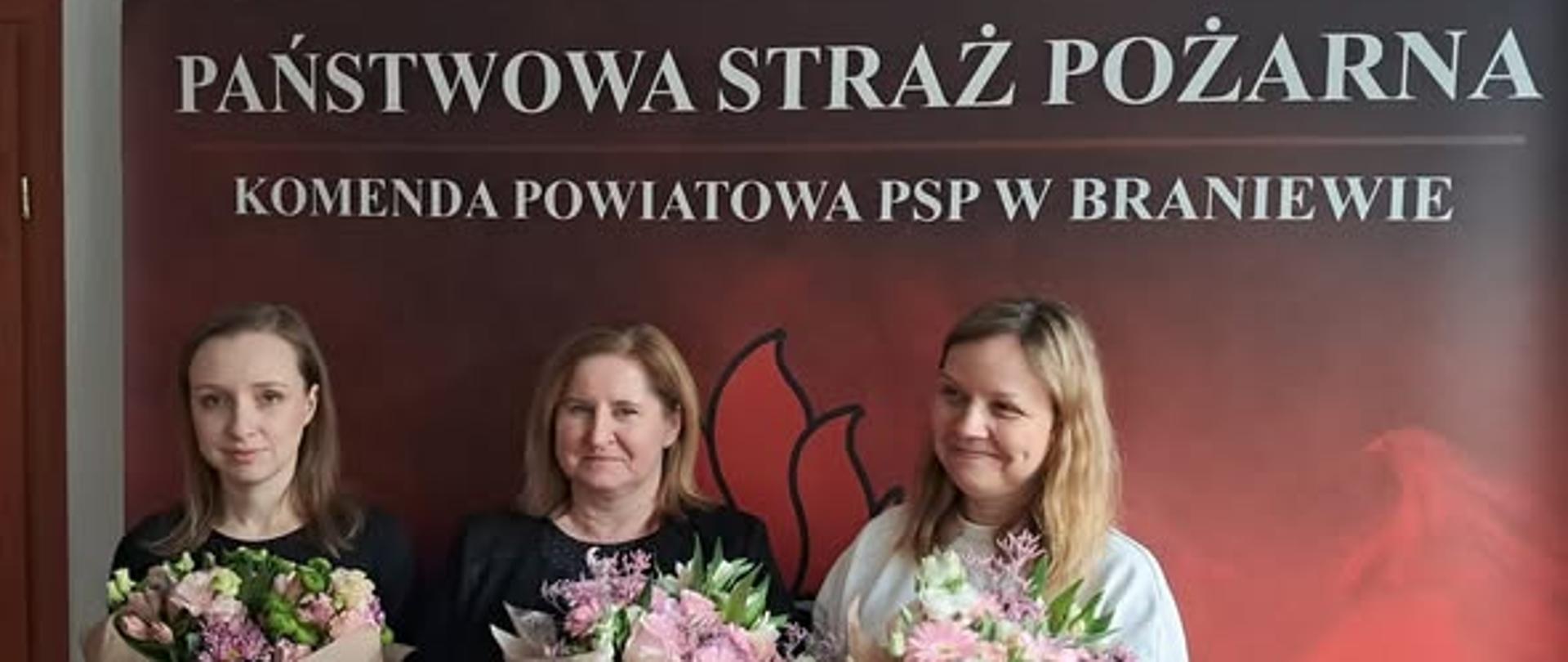 Na zdjęciu trzy kobiety z bukietami kwiatów w rekach. W tle baner z napisem państwowa straż pożarna komenda powiatowa PSP w Braniewie.