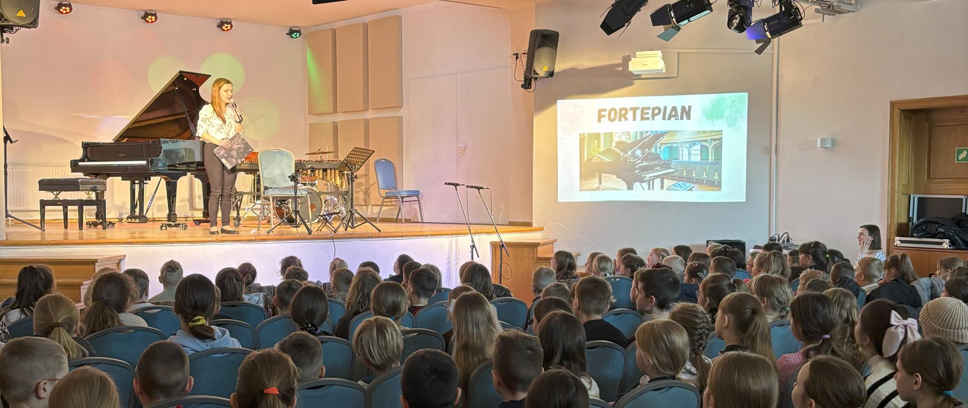 Na oświetlonej scenie stoi nauczycielka, która prowadzi koncert. Przed sceną widać licznie zgromadzoną publiczność. Po prawej stronie wyświetlany jest slajd z prezentacji multimedialnej. 