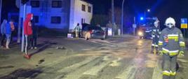 Widać dwóch strażaków, w oddali samochód straży pożarnej oraz uszkodzony samochód na skrzyżowaniu, jest noc