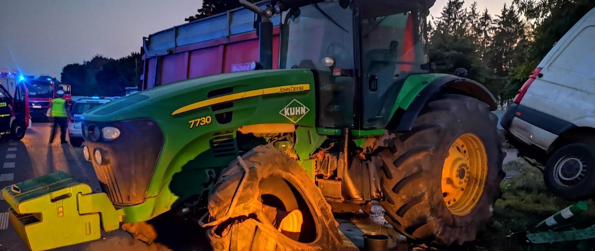 Zielony duży ciągnik rolniczy z uszkodzonym po zderzeniu z autem dostawczym lewym przednim kołem. W oddali przy radiowozie stoi policjant, dalej widać wozy strażackie.