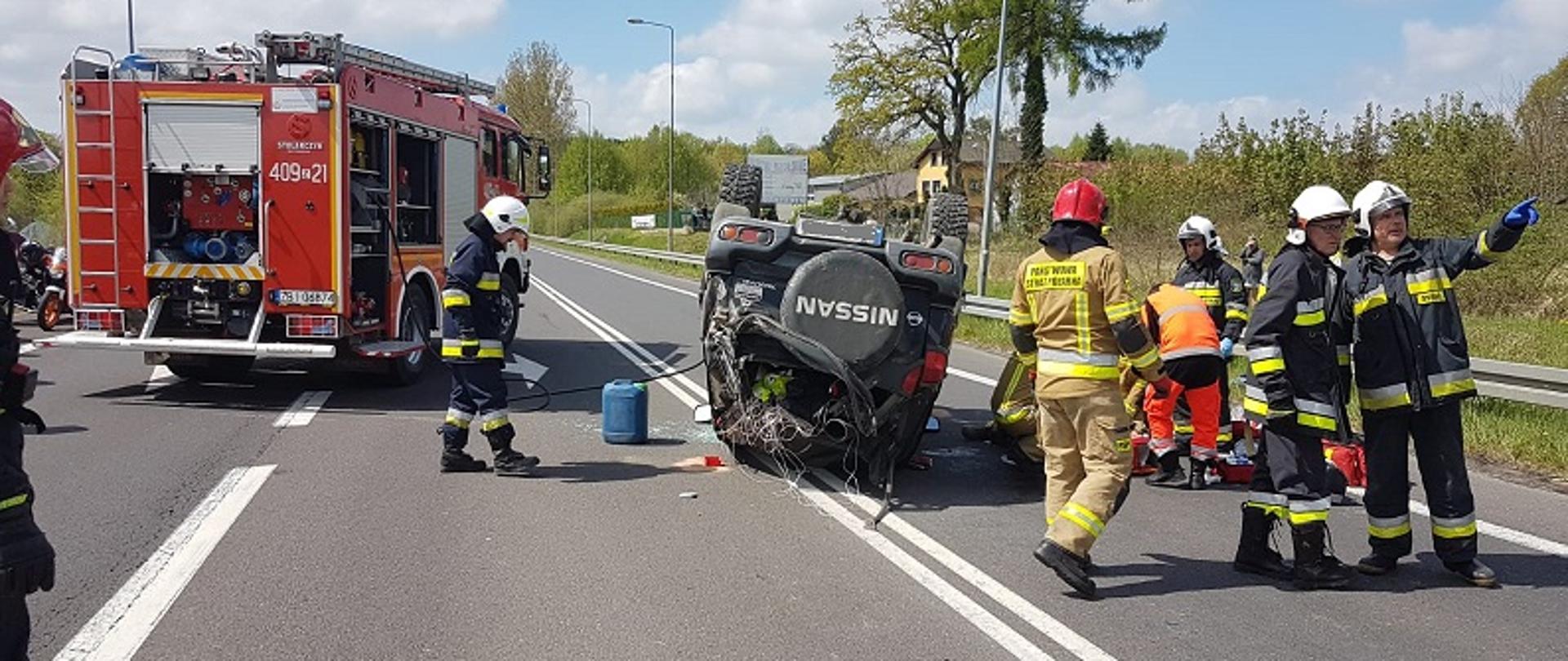 Zdjęcie przedstawia widoczny od tyłu, leżący na dachu szary samochód marki Nissan Terrano. Pojazd znajduje się na pasie ruchu, po prawej stronie strażacy w ubraniach specjalnych, po lewej czerwony samochód straży pożarnej