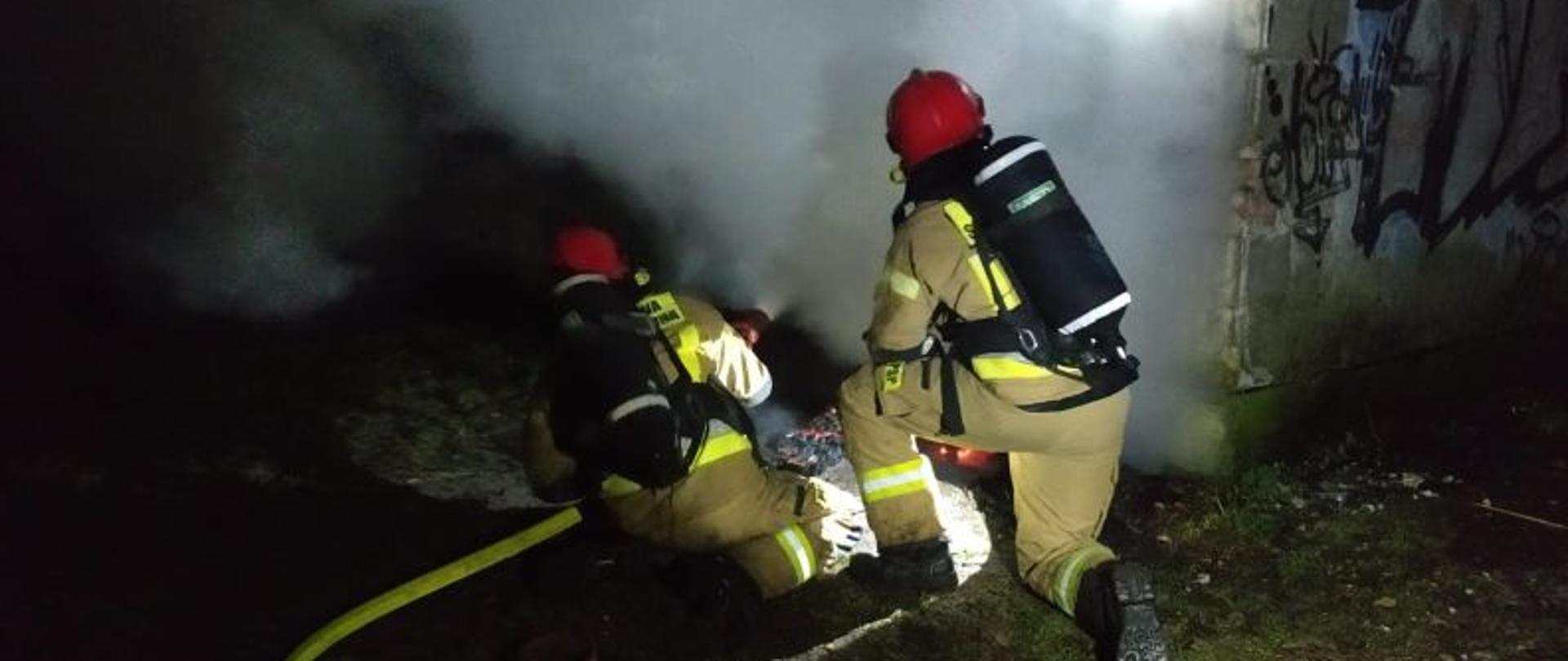 Zdjęcie wykonane w nocy. Dwóch strażaków przy garażu. Pochyleni przy wejściu do środka z którego wydostaje się dym i para powstała podczas gaszenia. Strażacy w piaskowych mundurach z linią gaśniczą w rękach. Strażacy mają na sobie aparaty ochrony dróg oddechowych