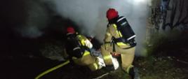 Zdjęcie wykonane w nocy. Dwóch strażaków przy garażu. Pochyleni przy wejściu do środka z którego wydostaje się dym i para powstała podczas gaszenia. Strażacy w piaskowych mundurach z linią gaśniczą w rękach. Strażacy mają na sobie aparaty ochrony dróg oddechowych