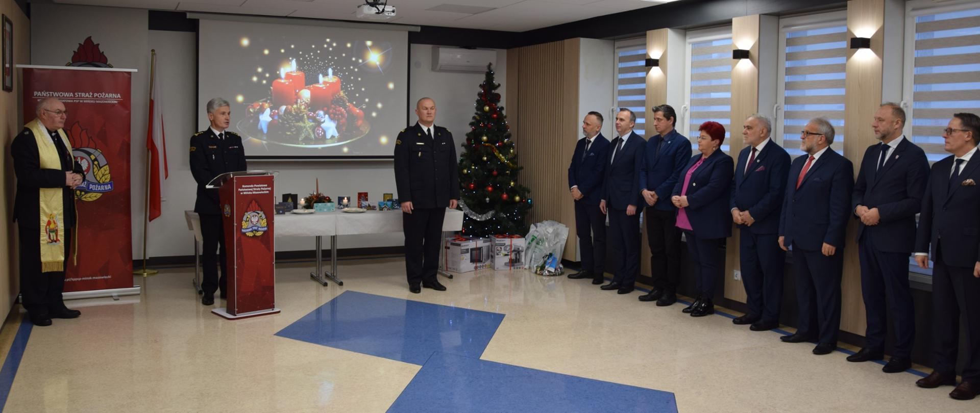 Komendant Powiatowy PSP w Mińsku Mazowieckim wita z mównicy wszystkich przybyłych na spotkanie. Na zdjęciu Kapelan Powiatowy Strażaków, Zastępca Komendanta PSP w Mińsku Mazowieckim oraz samorządowcy powiatu mińskiego i zaproszeni goście.