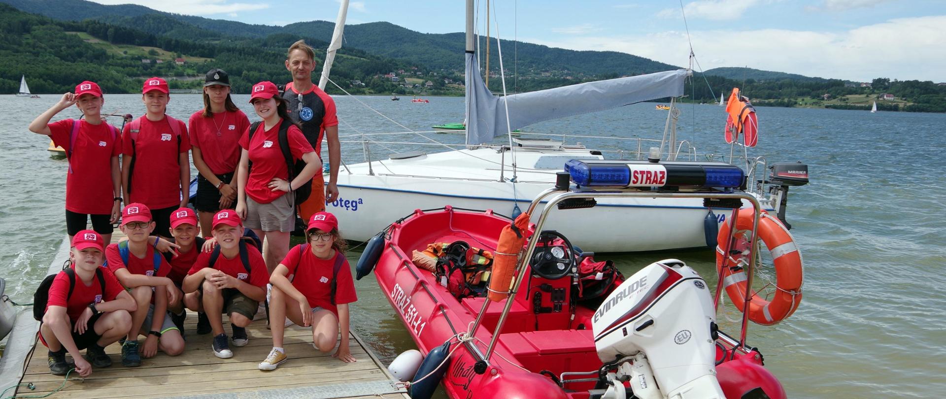 Widok z przodu. Drużyna MDP z ratownikiem wodnym na pomoście na jeziorze. W tle góry. Po prawej żaglówka oraz ponton strażacki z silnikiem zaburtowym. 
