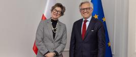 Kobieta i mężczyzna pozują na tle biało-czerwonej flagi Polski oraz flagi Unii Europejskiej. Uśmiechają się do kamery.