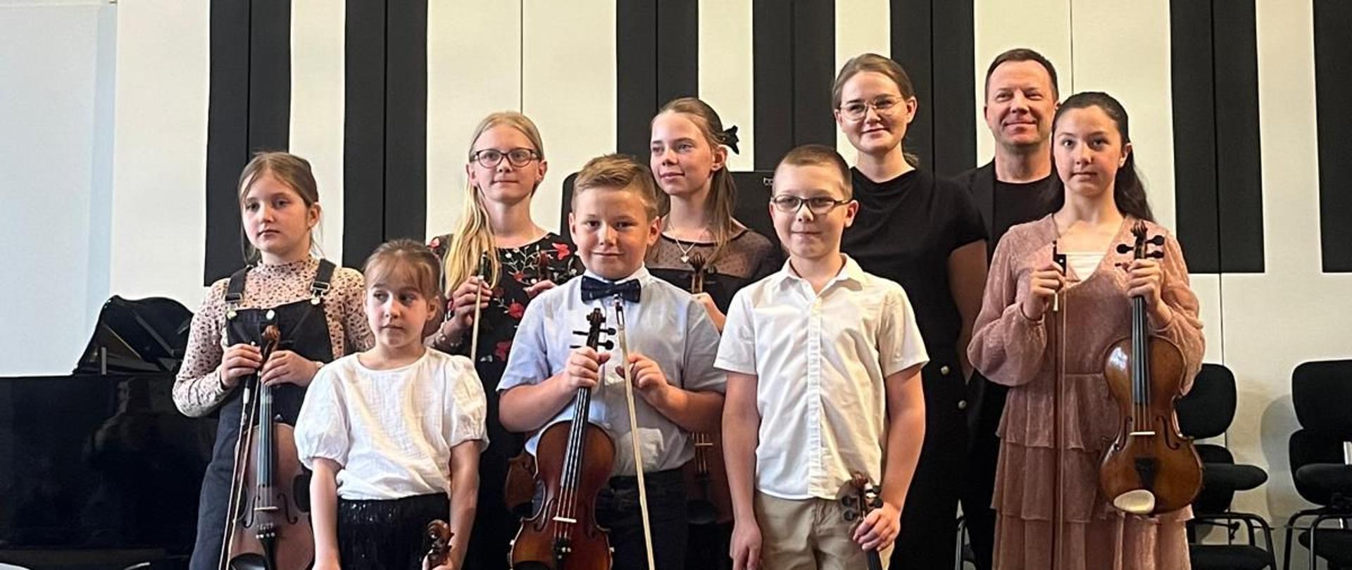 Kobieta, mężczyzna i wszyscy wykonawcy stoją na scenie trzymając skrzypce.