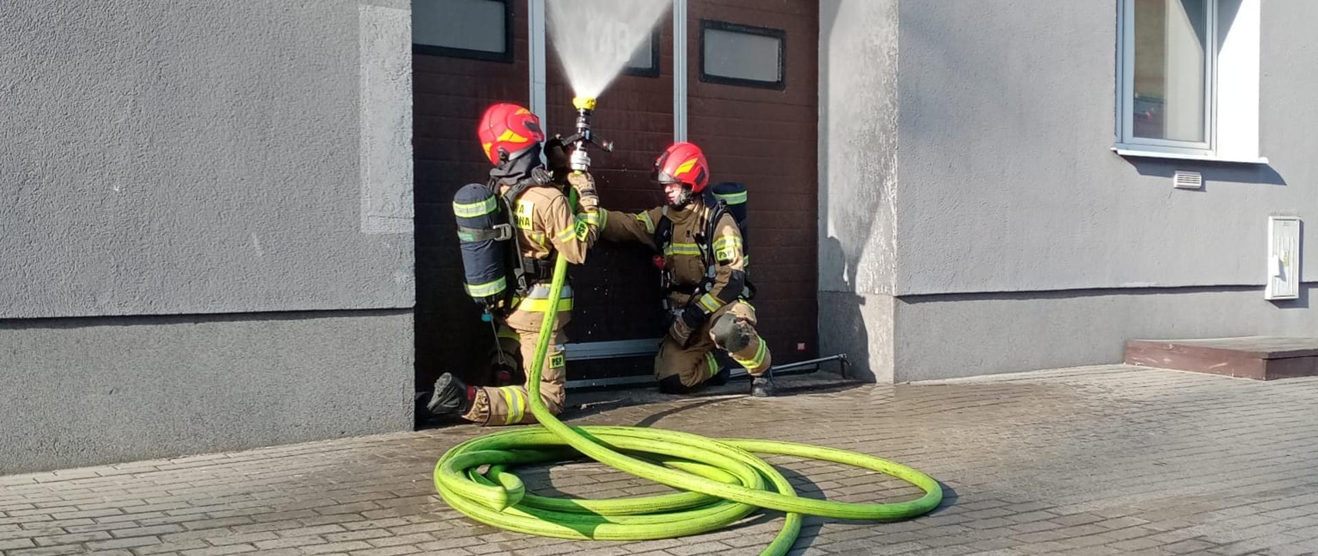 Przed wejściem do pomieszczenia garażowego, siedzi dwóch strażaków z zieloną linią wężową.